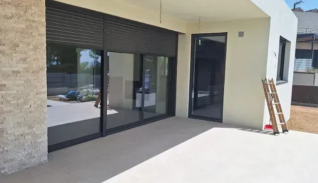 Exterior de un edificio moderno con grandes puertas corredizas de vidrio y un patio. Las persianas oscuras del lado izquierdo están cerradas.