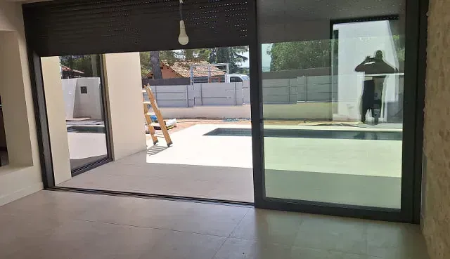 Vista interior de una casa moderna con grandes puertas corredizas de vidrio con vista a la piscina. Una persiana parcialmente abierta cuelga sobre la puerta izquierda.
