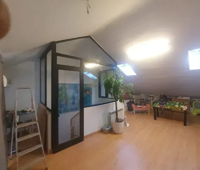 Una habitación abuhardillada con una mampara de cristal con marco negro, suelo de madera y una maceta. También hay una escalera de mano y muebles.