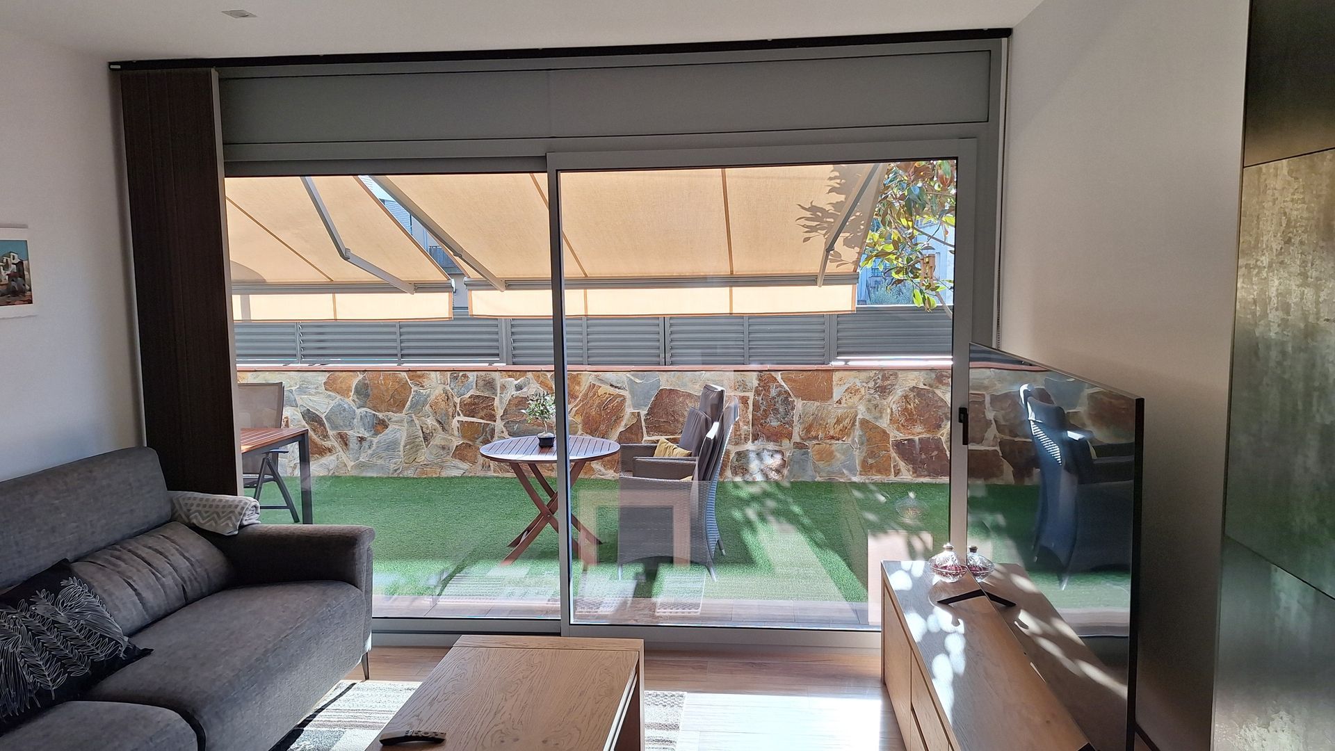 Sala de estar con puertas corredizas de vidrio que dan a un patio, sofá gris, mesa de centro de madera y un televisor colgado en la pared. El patio tiene una mesa pequeña, una parrilla y paredes de piedra.