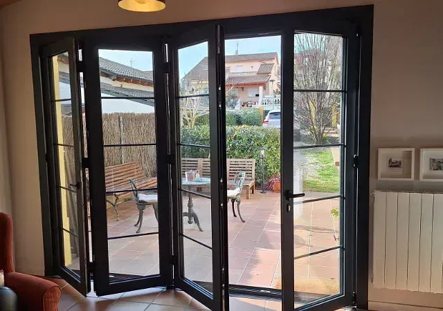 Las puertas de vidrio plegables con marco negro se abren a un patio con asientos al aire libre, con vista a un jardín.