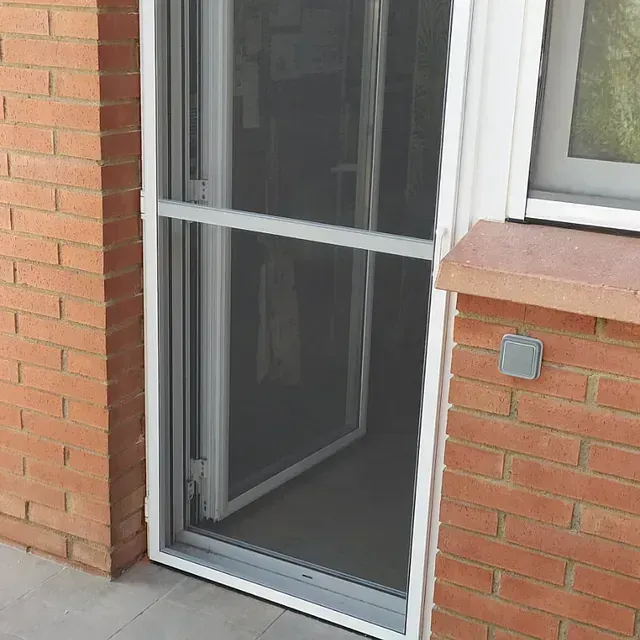 Una puerta mosquitera retráctil con marco blanco en un edificio de ladrillo, abierta a una habitación.