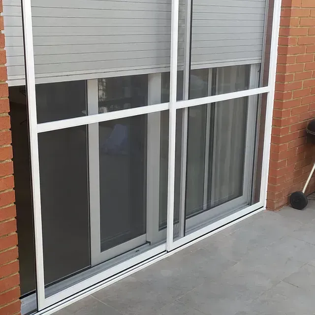 Puerta corredera blanca con mosquitera frente a una ventana cerrada con persianas grises. Pared de ladrillo a la derecha.