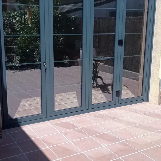Un conjunto de puertas de vidrio con marco verde azulado refleja un patio con baldosas de terracota y una mesa, situado en un día soleado.