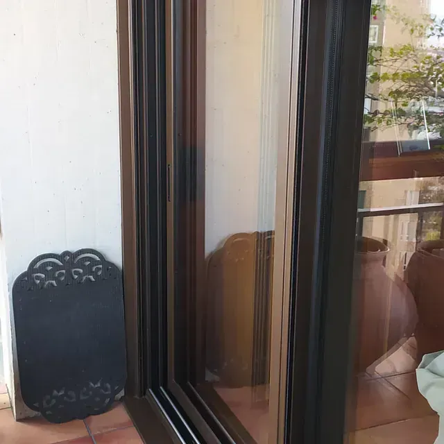 Una puerta corrediza de vidrio con marco oscuro que refleja el entorno exterior, junto a una pizarra decorativa en un patio con macetas de terracota.