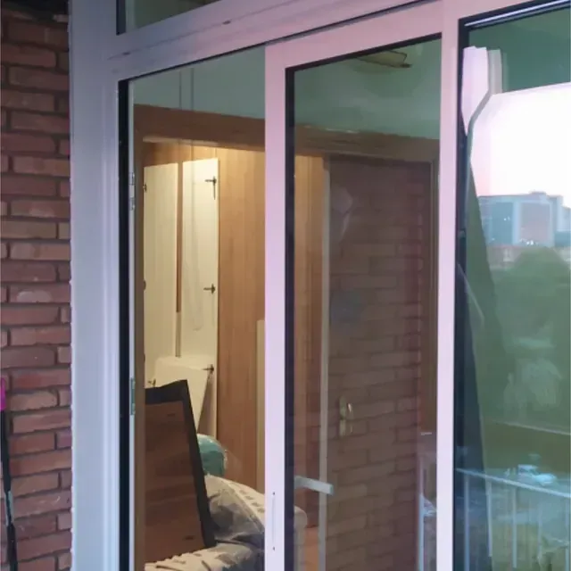 Puerta corrediza de vidrio con marco blanco, que revela un interior poco iluminado con un vistazo a un pasillo, puertas y muebles.