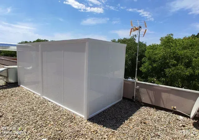 Estructura rectangular blanca sobre una azotea, con una antena y árboles al fondo bajo un cielo azul.