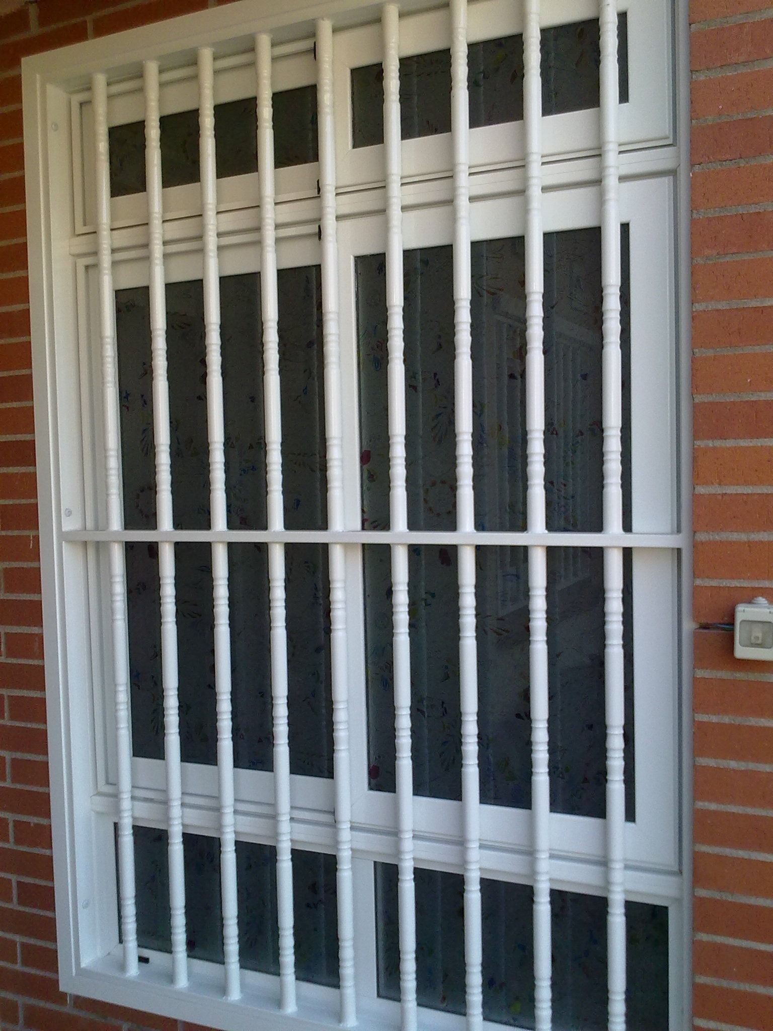 Ventana blanca con rejas de seguridad, ubicada en un edificio de ladrillo rojo. Las rejas, verticales y horizontales, cubren el cristal.