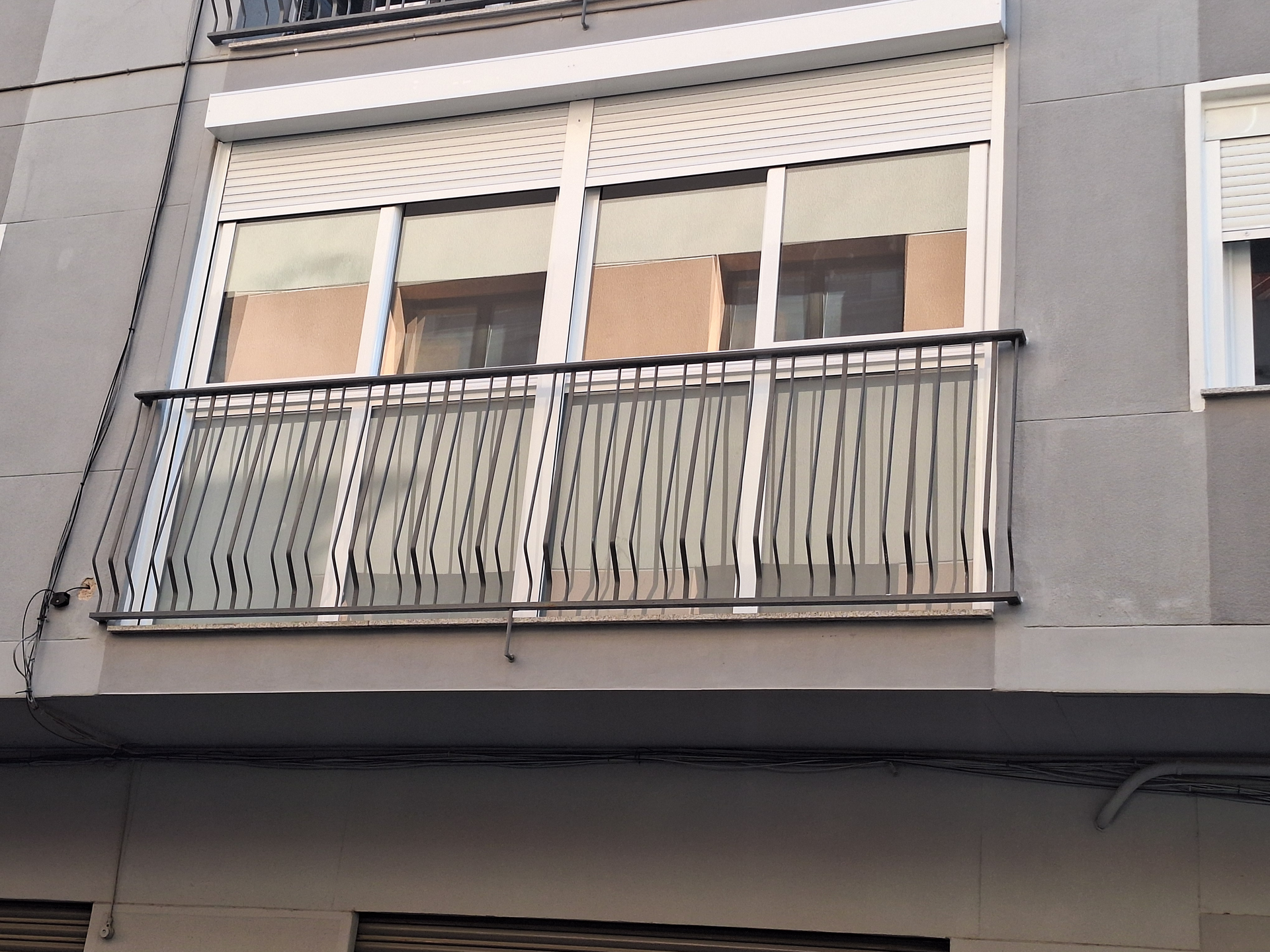 Primer plano de la fachada de un edificio con balcón y ventanas. Exterior gris, marcos de ventanas blancos, persianas cerradas y barandilla negra.
