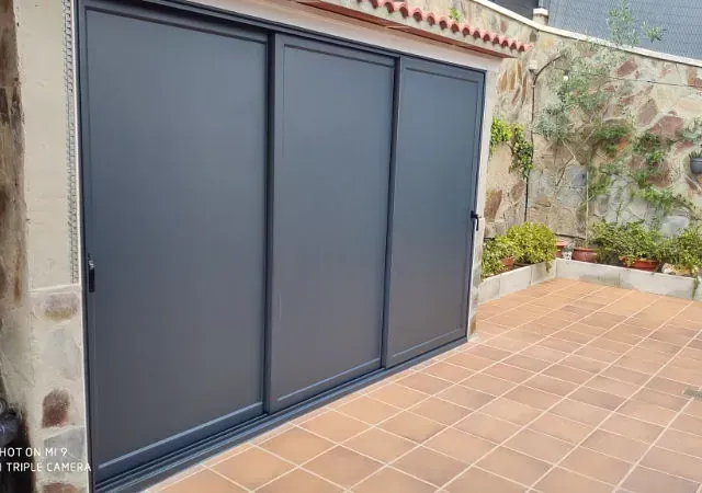 Tres puertas correderas gris oscuro dan a un patio con baldosas de terracota. Al fondo, un muro de piedra y vegetación.