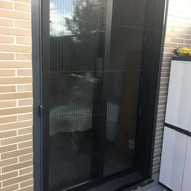 Una puerta mosquitera retráctil negra sobre una pared de ladrillos, con una unidad de almacenamiento blanca y algunas herramientas visibles.