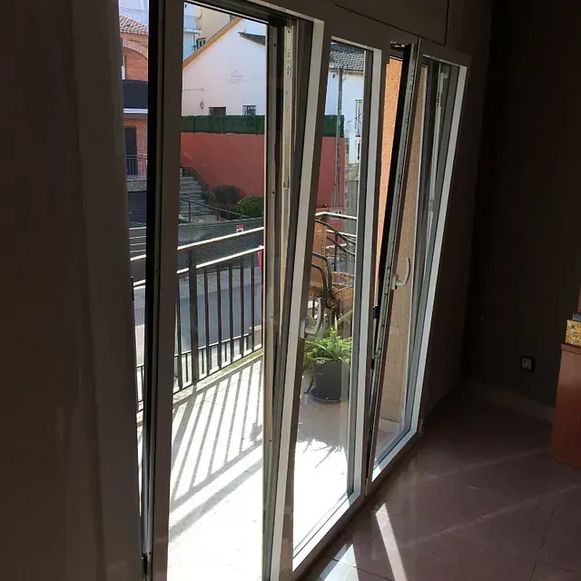 Vista interior de una puerta de balcón de cristal parcialmente abierta con barandilla negra y una planta en maceta visible desde el exterior. La luz del sol entra a raudales.