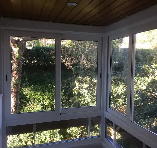 Terraza acristalada esquinera con ventanas de marco blanco y vistas a la frondosa vegetación. Techo revestido de madera.