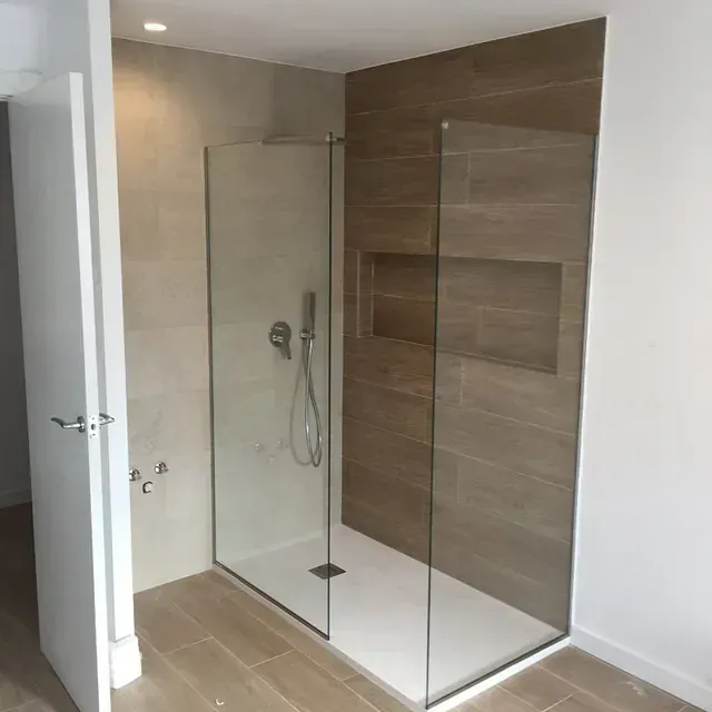 Un baño moderno con ducha de cristal. Se aprecian paredes de azulejos marrones y beige, un plato de ducha blanco y una puerta blanca parcialmente abierta.