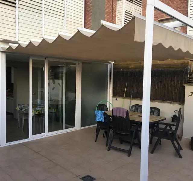 Un patio con toldo beige, puertas corredizas de cristal, una mesa y sillas negras. El edificio es residencial.