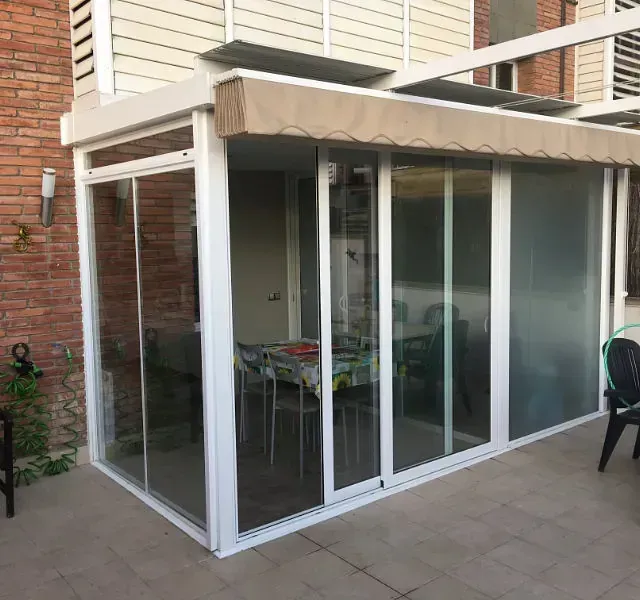 Patio cerrado con marcos blancos, puertas corredizas de vidrio y un toldo beige. Se ve una mesa de comedor en el interior.