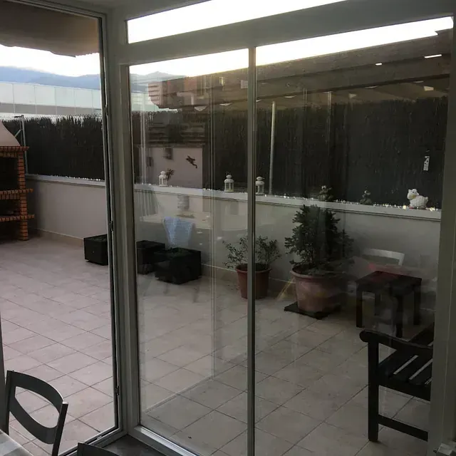 Terraza con paredes de cristal, suelo de baldosas, plantas en macetas y una parrilla de ladrillo al fondo.
