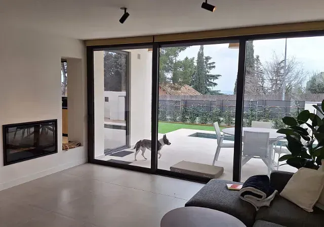 Vista interior de una moderna sala de estar con grandes puertas corredizas de vidrio que dan a un patio trasero. Un perro está de pie cerca de la puerta abierta.