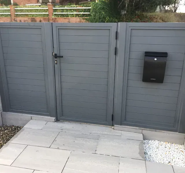 Sección de portón y valla gris con buzón negro. El portón tiene manija y bisagras. Camino pavimentado al frente.