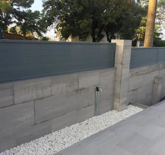 Un muro de hormigón gris rematado con una valla gris a juego. Hay un grifo de agua en la pared. Piedras blancas bordean la base.
