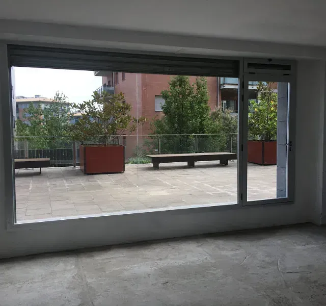 Vista desde una habitación con un gran ventanal que da a un patio con árboles en grandes jardineras, bancos y edificios.