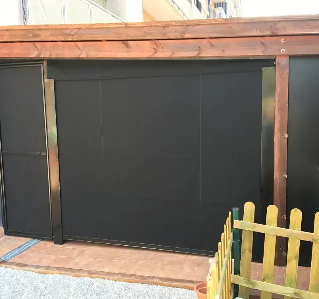 Un parasol negro cubre una zona exterior, enmarcada por vigas de madera y metal. Una pequeña valla se alza en primer plano.