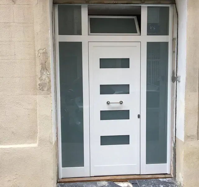 Puerta blanca con paneles de vidrio esmerilado a ambos lados y en la parte superior. Tiene un tirador plateado horizontal y cuatro ventanas rectangulares. Está ubicada en un edificio beige.