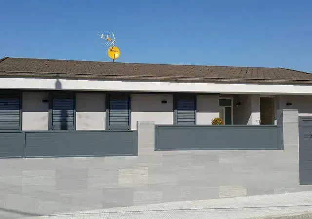 Una casa de un piso con una pared de piedra gris, una cerca gris y una antena de televisión en el techo.