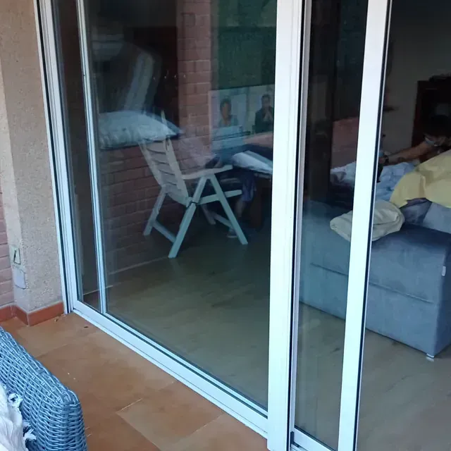 Una puerta corrediza de vidrio blanca refleja un patio y un interior con una persona en un sofá y una silla.