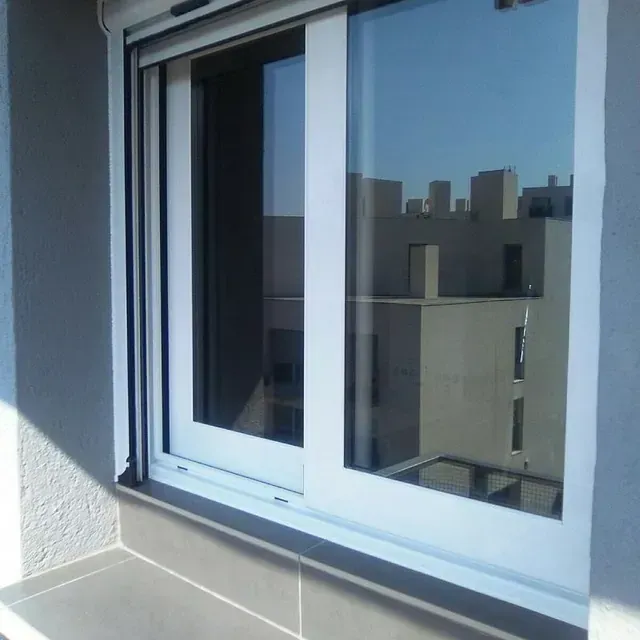 Ventana corrediza con marco blanco que refleja los edificios y el cielo azul.