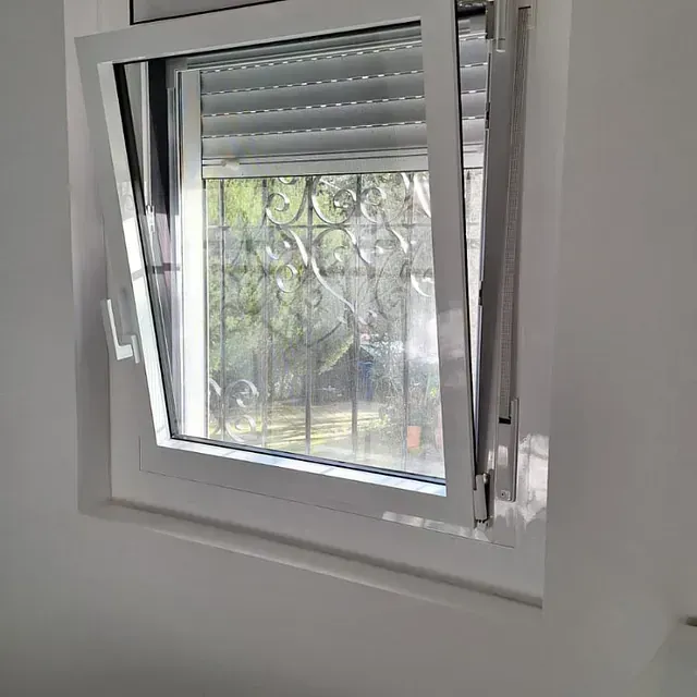 Ventana abatible blanca, parcialmente abierta, que revela una vista de un jardín a través de rejas de seguridad y una persiana cerrada.