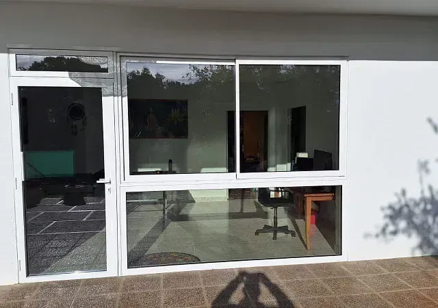 Vista exterior de una puerta corrediza de vidrio con marco blanco y una puerta lateral. La luz del sol se refleja en el vidrio, mostrando un espacio de oficina interior.