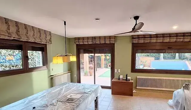 Una habitación con paredes verdes, marcos de ventanas marrones y una puerta corrediza que da a un patio trasero. Una mesa está cubierta de plástico y se ve un ventilador de techo.