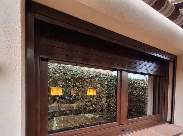 Ventana marrón con contraventana a juego parcialmente cerrada que refleja un jardín con lámparas.