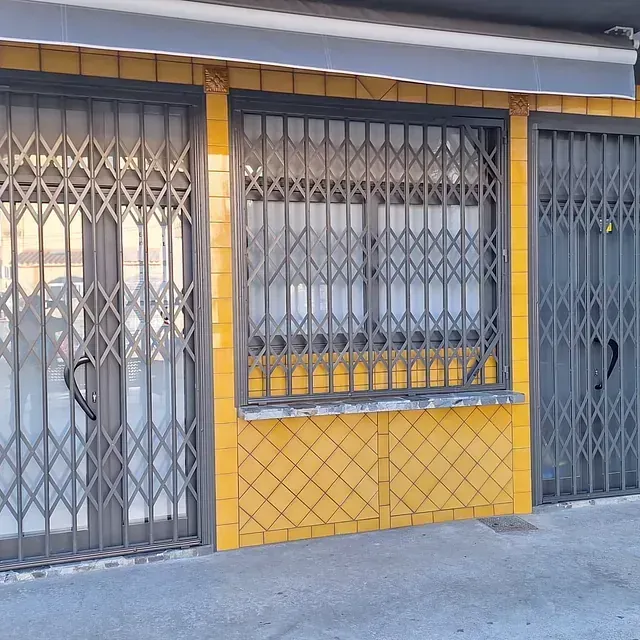 Una tienda con paredes de azulejos amarillos, una ventana y puertas, todo ello protegido por rejas metálicas grises. Un toldo gris corona el edificio.