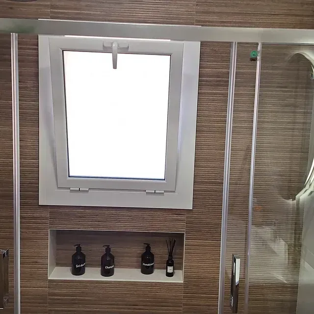 Baño con ventana con marco blanco, ducha de cristal y estante empotrado con artículos de tocador en una pared de azulejos marrones.