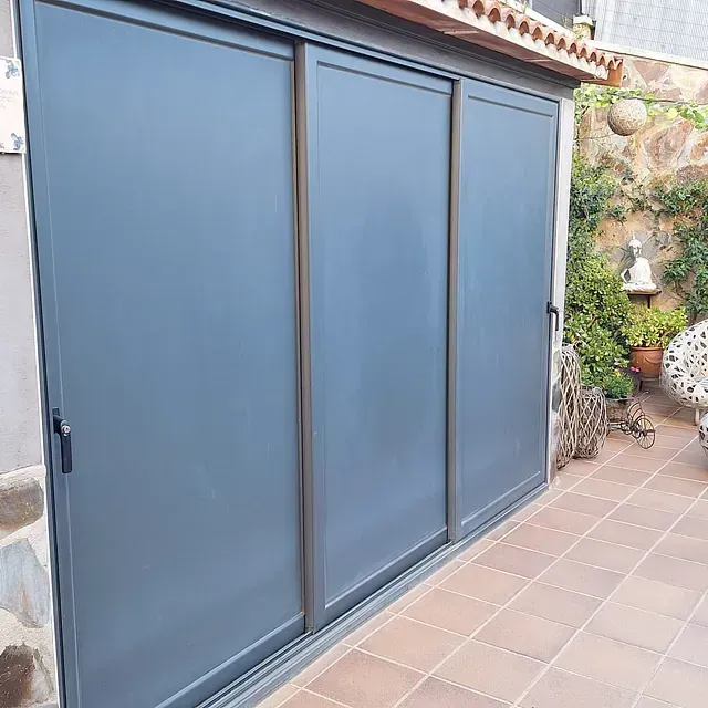Tres puertas corredizas grises en un patio de baldosas, con un fondo de vegetación y una pared de piedra.
