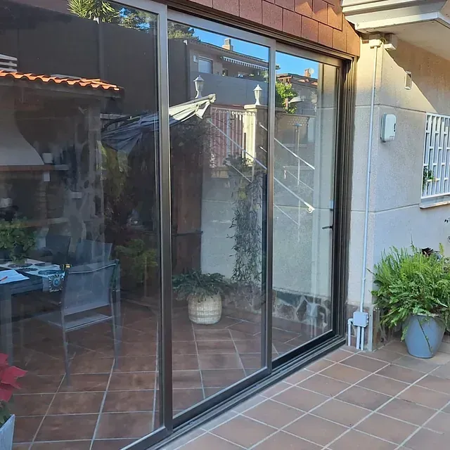 Unas puertas corredizas de vidrio dan acceso a un patio de baldosas con plantas en macetas. En el cristal se reflejan un patio trasero y una escalera.