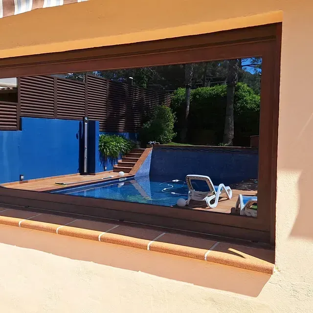 Vista de una piscina desde una ventana. La piscina está rodeada de paredes azules y una terraza de madera, con tumbonas y un fondo arbolado.