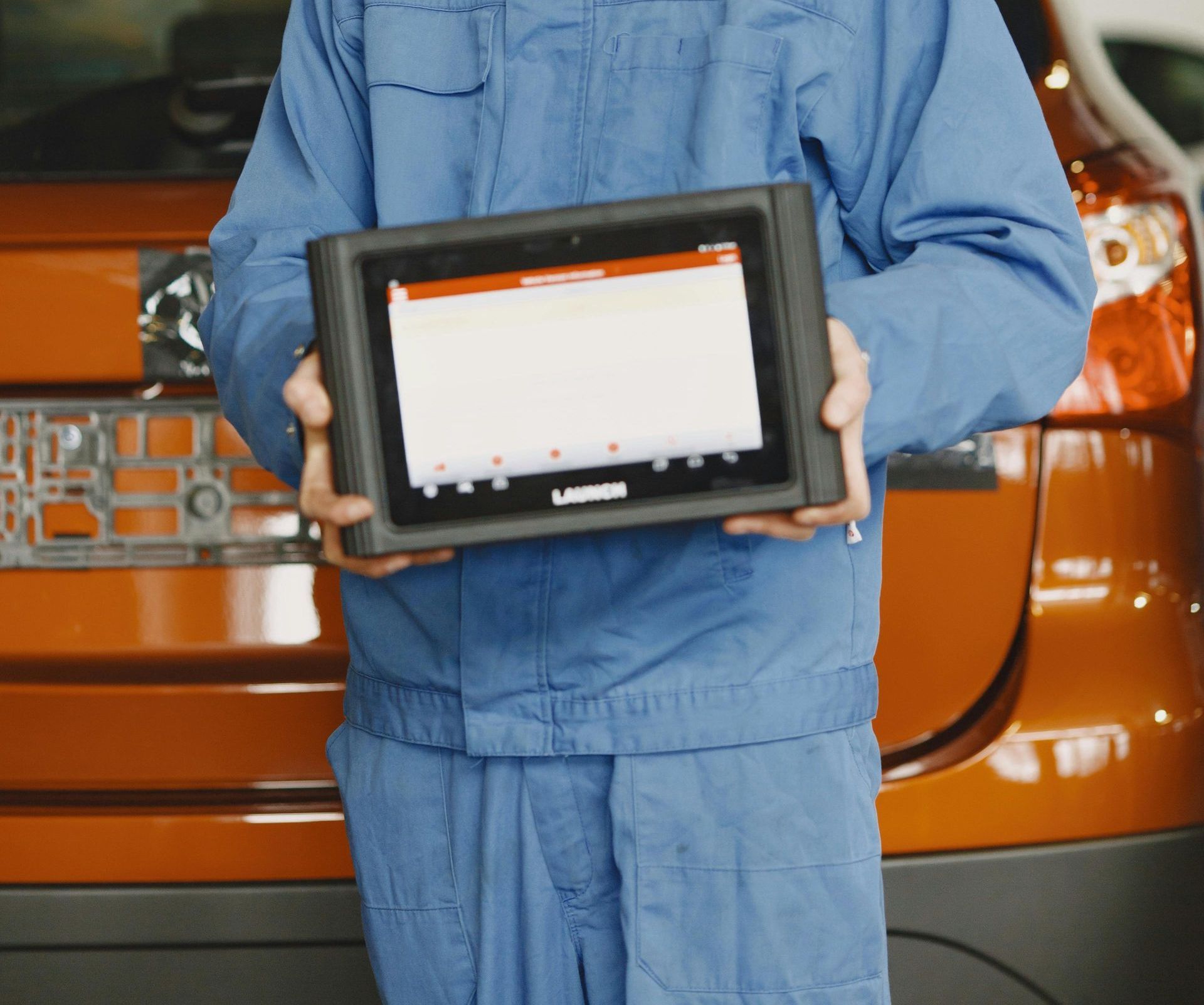 Un hombre con un mono azul sostiene una tableta delante de un coche naranja.
