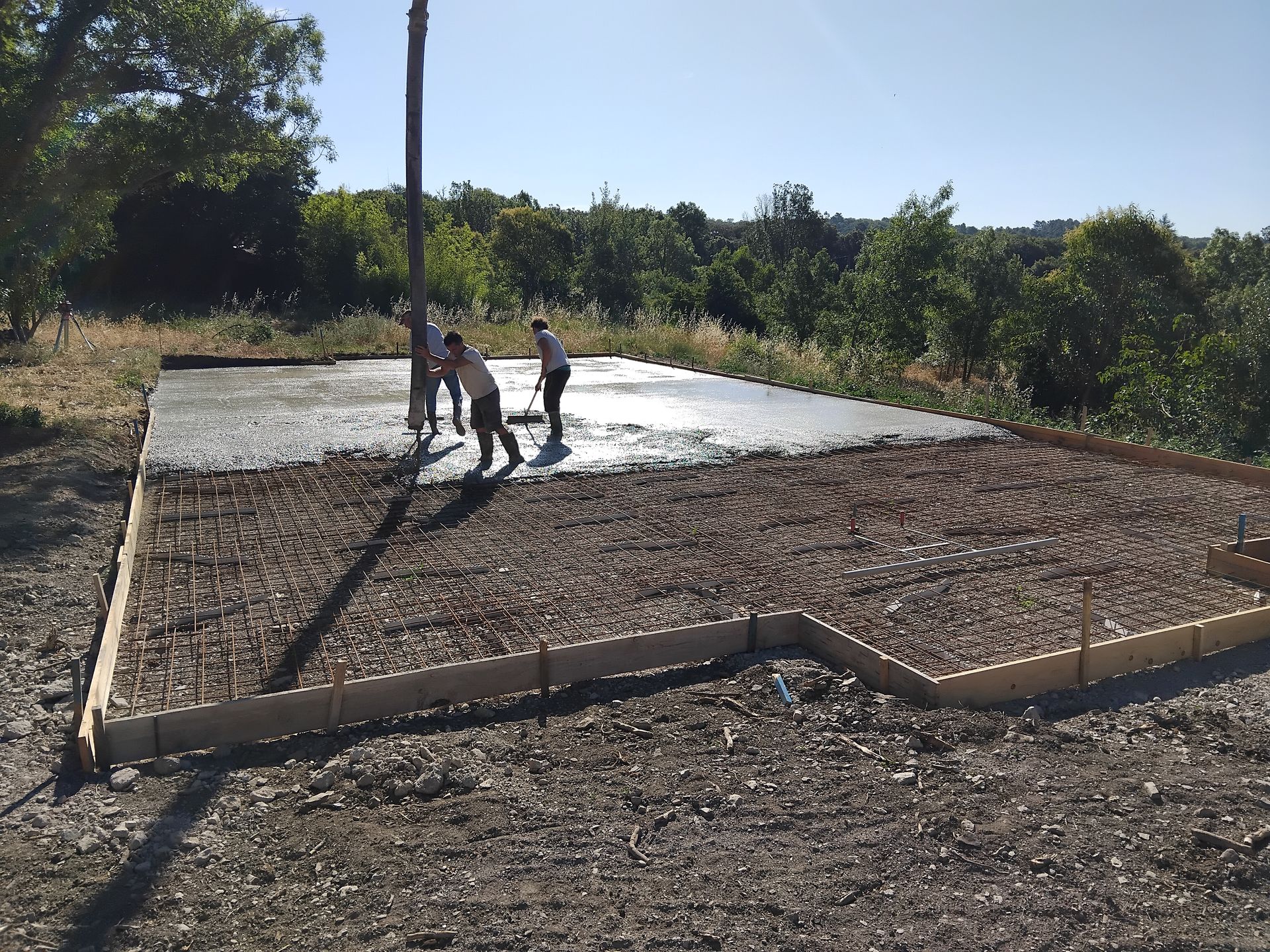 Des ouvriers coulent du béton sur des fondations renforcées d'armatures. On aperçoit des coffrages en bois, des arbres et le ciel bleu.