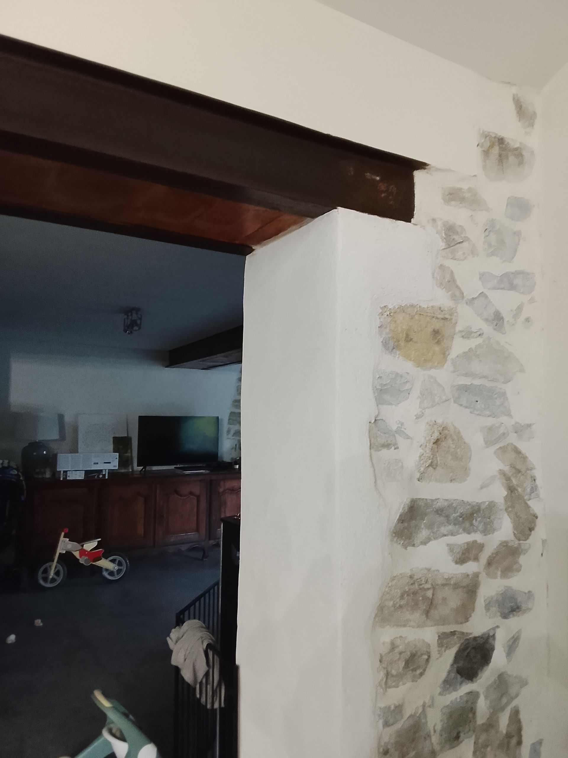 Mur en pierre et plâtre encadrant une porte, avec vue sur un salon et un jouet d'enfant.