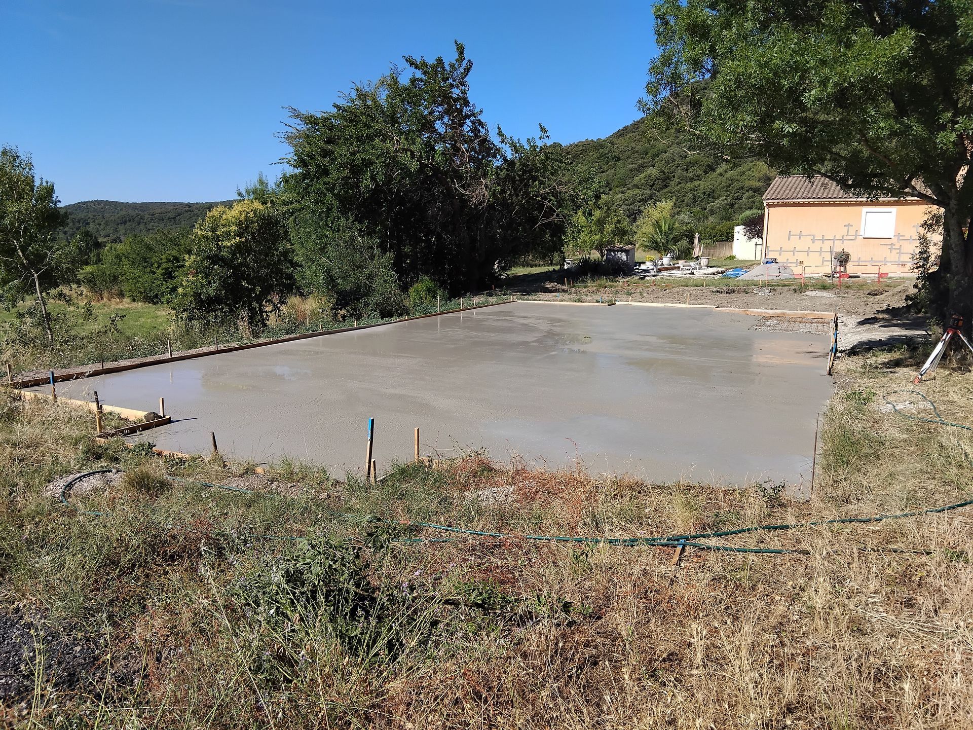 Fondations en béton fraîchement coulées dans une cour gazonnée, entourées d'une charpente en bois, avec une maison et des arbres en arrière-plan.