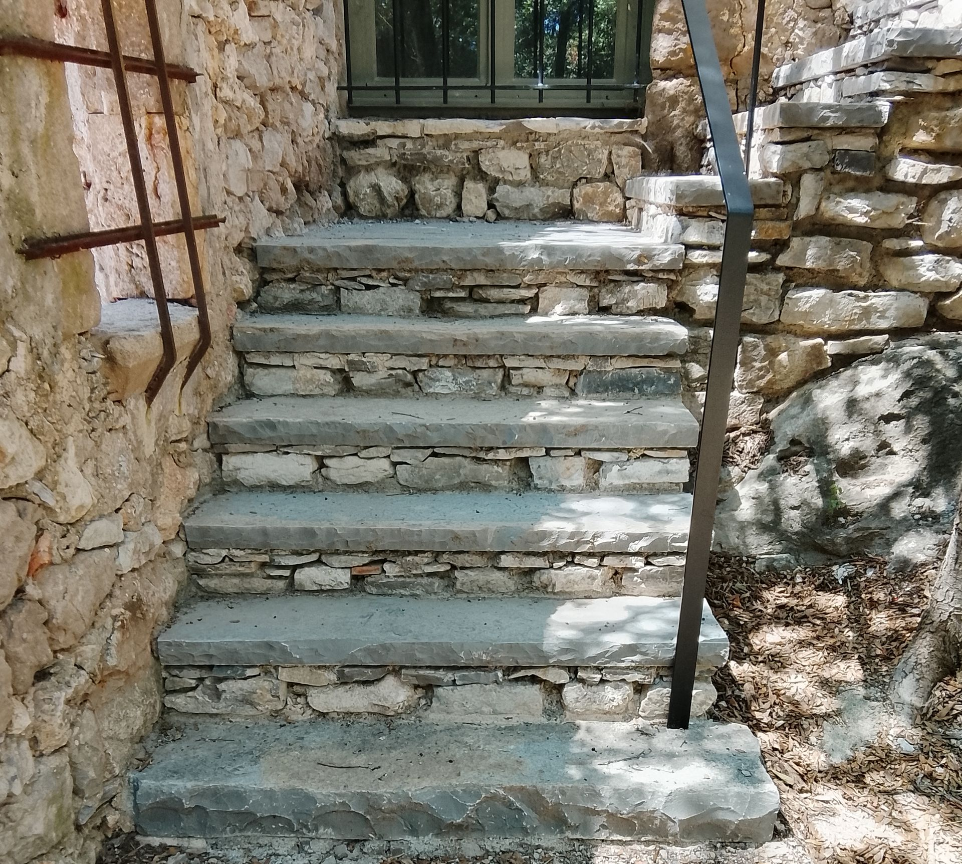 Des marches en pierre mènent à une fenêtre aménagée dans un mur en pierre brute. Une rampe métallique est présente.