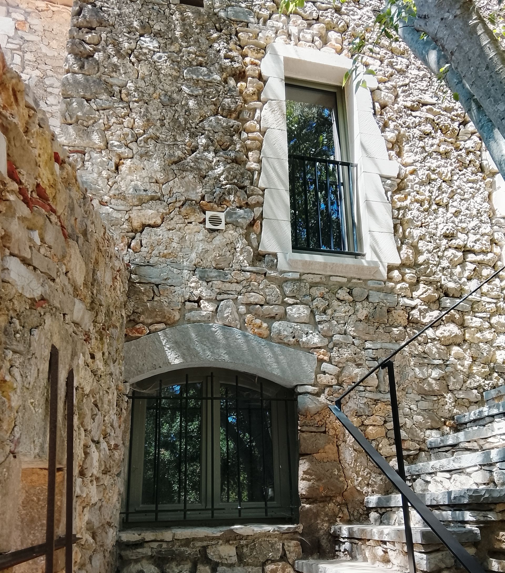 Façade en pierre d'un bâtiment avec deux fenêtres. La fenêtre du haut est munie d'une rambarde noire. Escalier extérieur à droite.