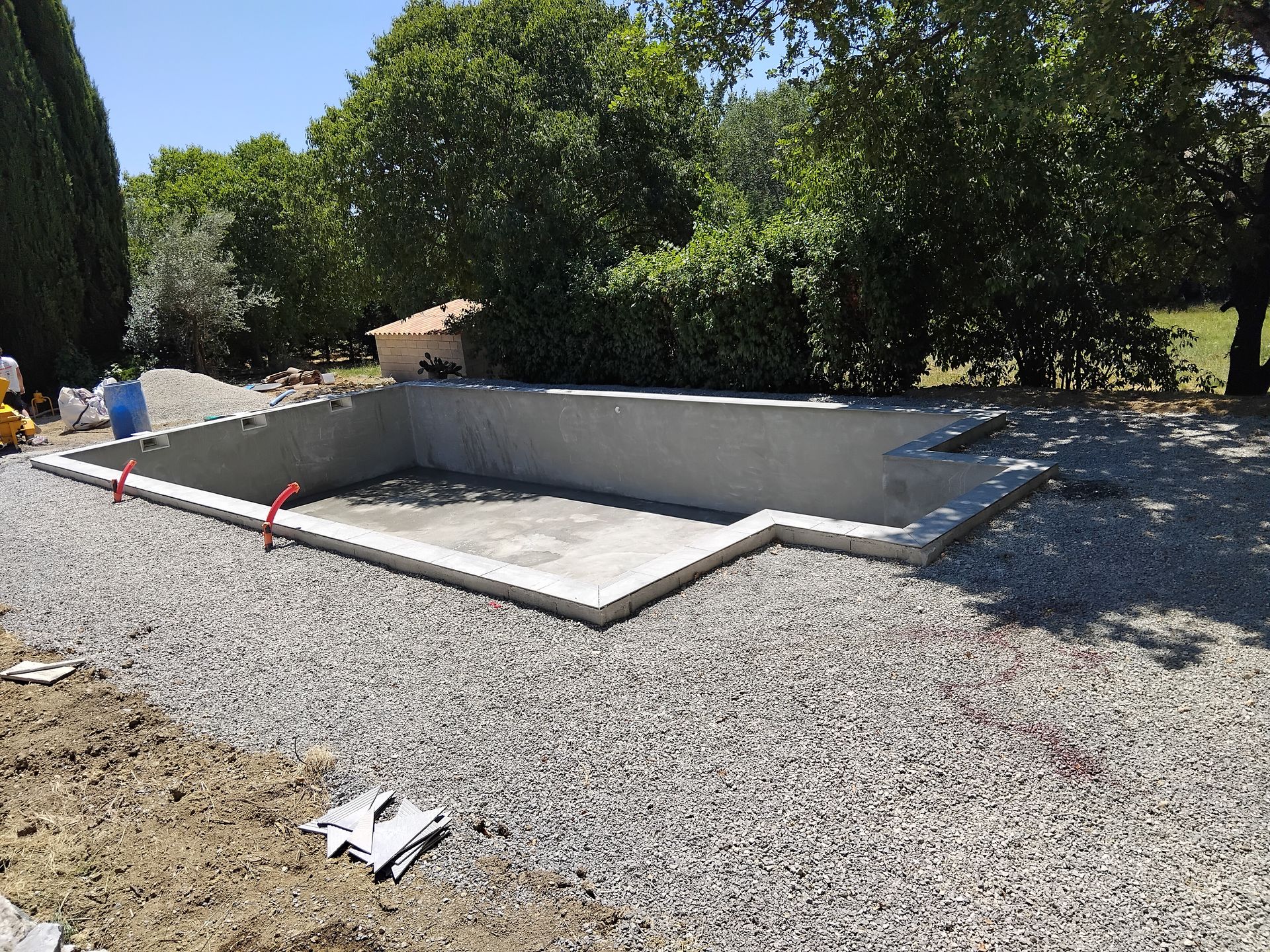 Piscine en béton en construction dans une cour gravillonnée, avec des arbres en arrière-plan.