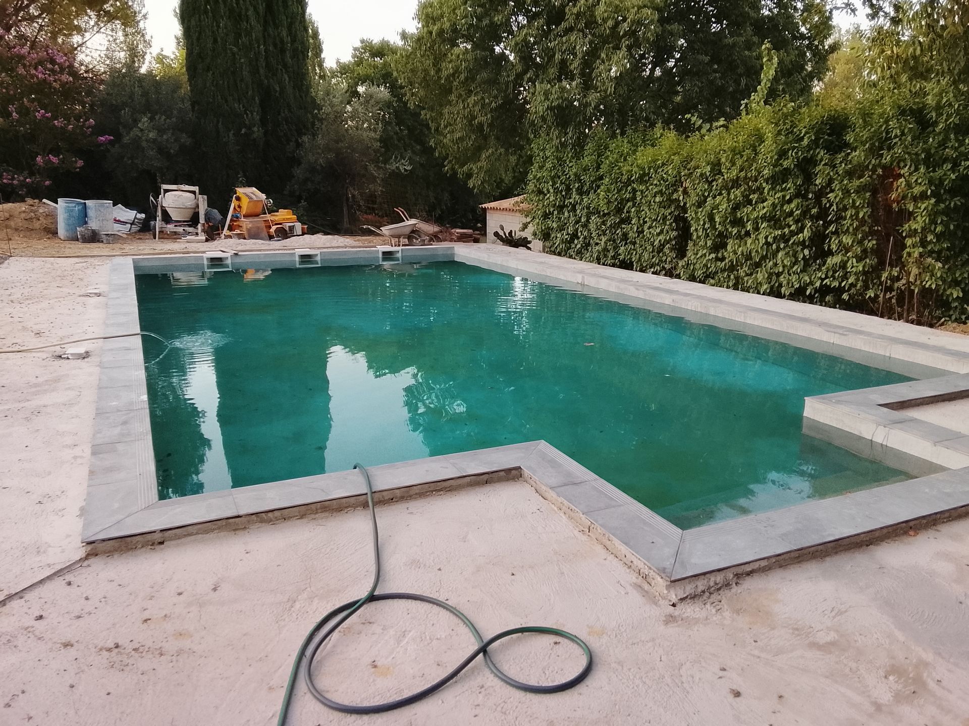 Piscine rectangulaire remplie d'eau turquoise ; bordure en pierre grise, entourée de béton, d'arbres et d'engins de chantier.