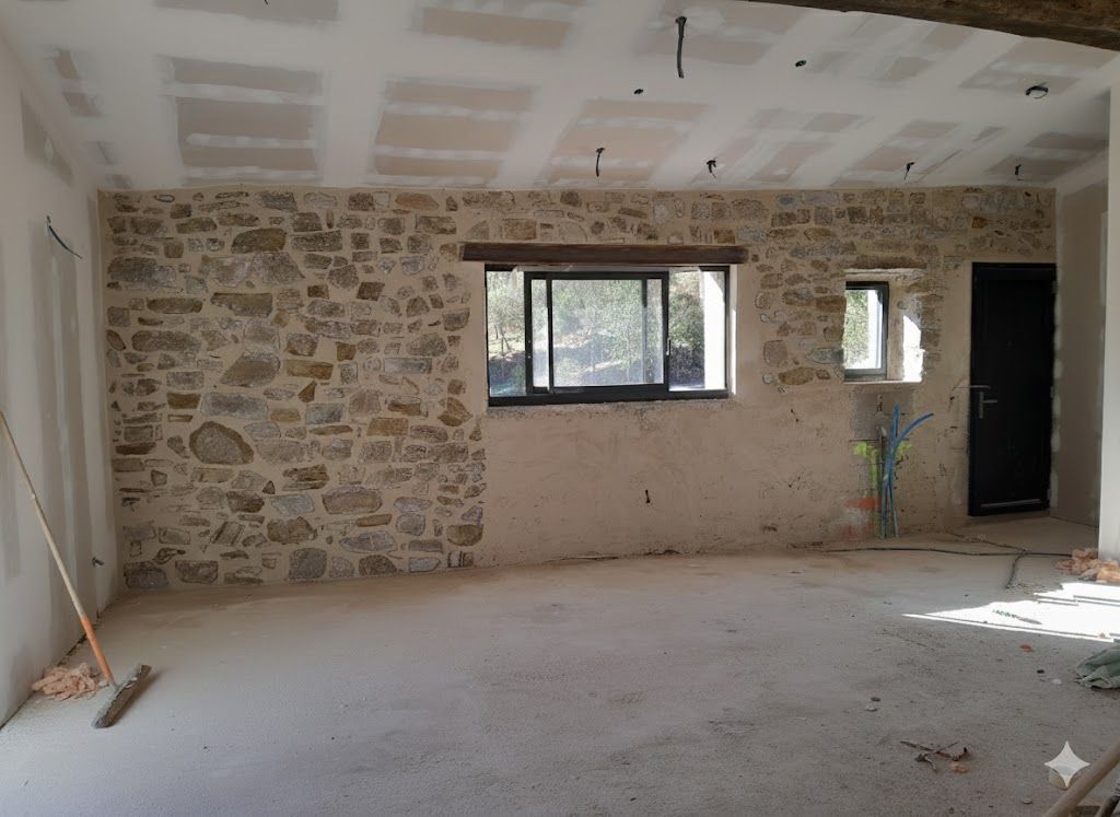 Pièce avec murs en pierre apparente, fenêtres, plafond brut et sol en béton.