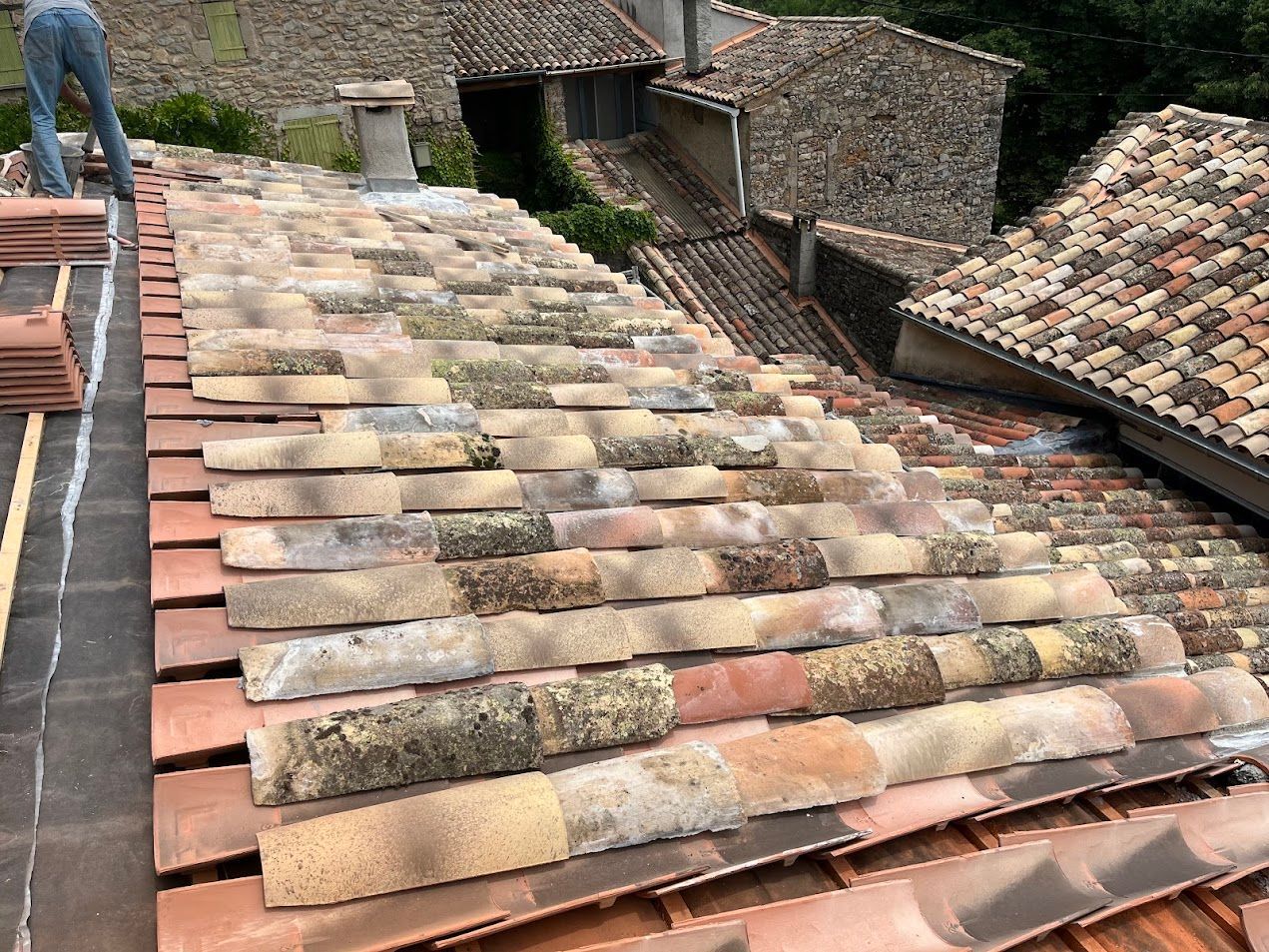 Un couvreur pose des tuiles en terre cuite sur un toit abîmé par les intempéries. On distingue des tuiles neuves et anciennes, avec une personne en arrière-plan.