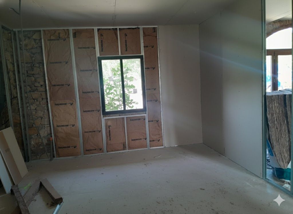 Pièce intérieure en travaux, avec isolation apparente dans un mur et une fenêtre. Plaque de plâtre installée sur l'autre mur.
